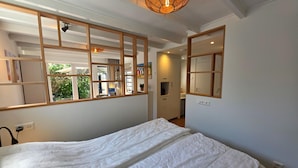 A bedroom with a large bed, a window with a view of greenery, and a ceiling light fixture. - Charming and Cozy Furnished 2-person Studio Apartment (De Koog)