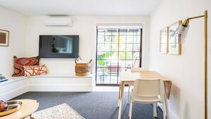 A modern room with a flat-screen TV mounted on the wall, a dining table with chairs, a small seating area with cushions, and a large window with a view of the outdoors. - Charming 1-bedroom cottage near SF (Sausalito)