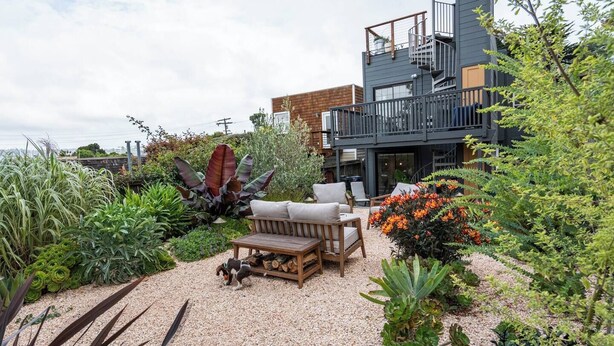 A modern two-story building with a balcony, surrounded by a garden with various plants and a seating area with a table and chairs. - Charming 1-bedroom cottage near SF (Sausalito)