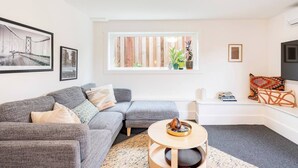 A modern living room with a grey sofa, a small round table, and a window with wooden blinds. - Charming 1-bedroom cottage near SF (Sausalito)