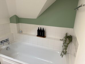 Une salle de bain avec une baignoire blanche, un mur vert, des carreaux blancs, une étagère avec des bouteilles noires et une plante.