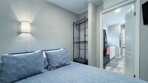 Quarto com uma cama, uma luminária na parede, uma estante de metal e uma porta que dá para o banheiro.