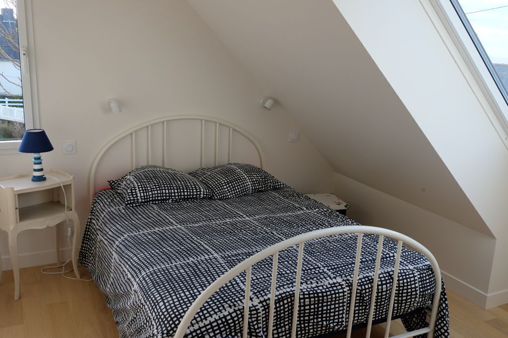 Une chambre avec un plafond en pente, un cadre de lit en métal blanc, une couvre-lit à motifs, une table de chevet avec une lampe et une fenêtre donnant sur l’extérieur.