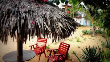 Un paraguas de paja con dos sillas rojas debajo, una zona arenosa y algo de vegetación.