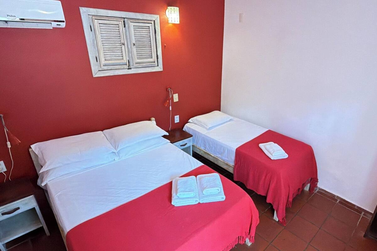Habitación de hotel con dos camas, cada una con sábanas blancas y colchas rojas. Se observa un aire acondicionado instalado en la pared sobre las camas, una mesita de noche con lámpara y un armario a la derecha.