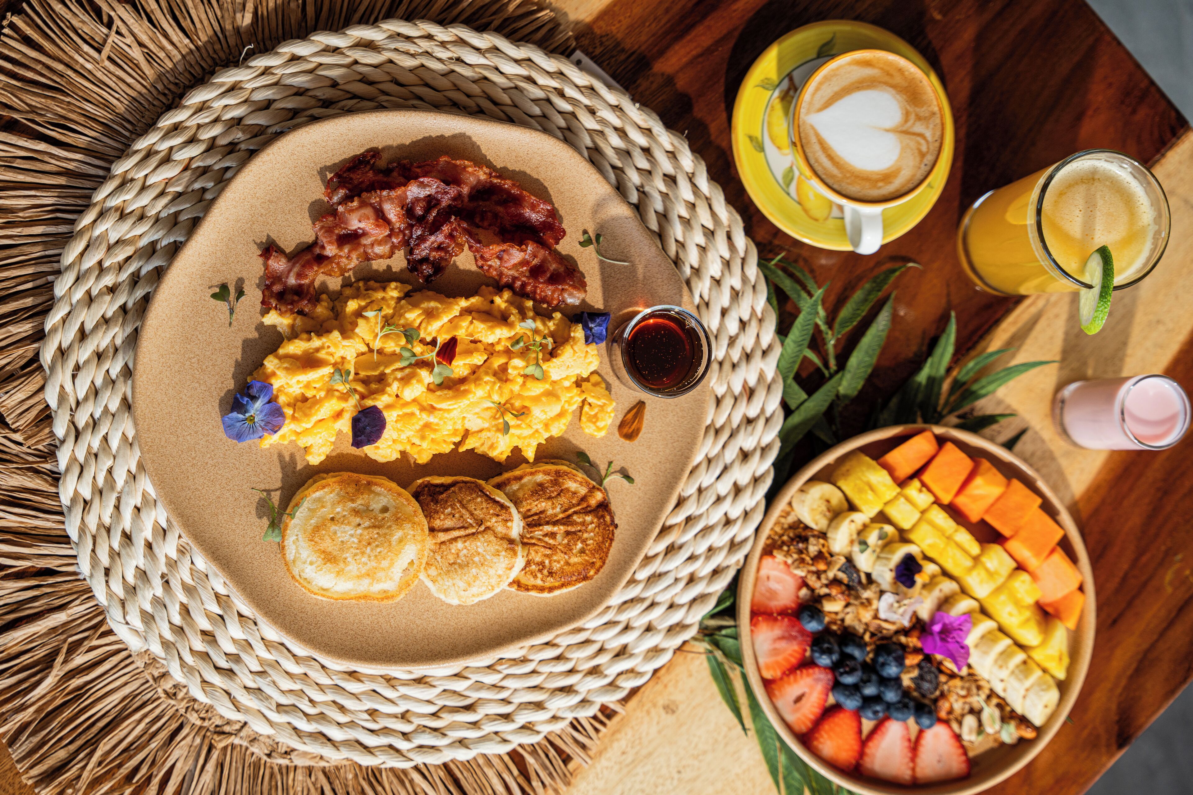 A breakfast setting with scrambled eggs, bacon, pancakes, and a fruit bowl on a woven mat.