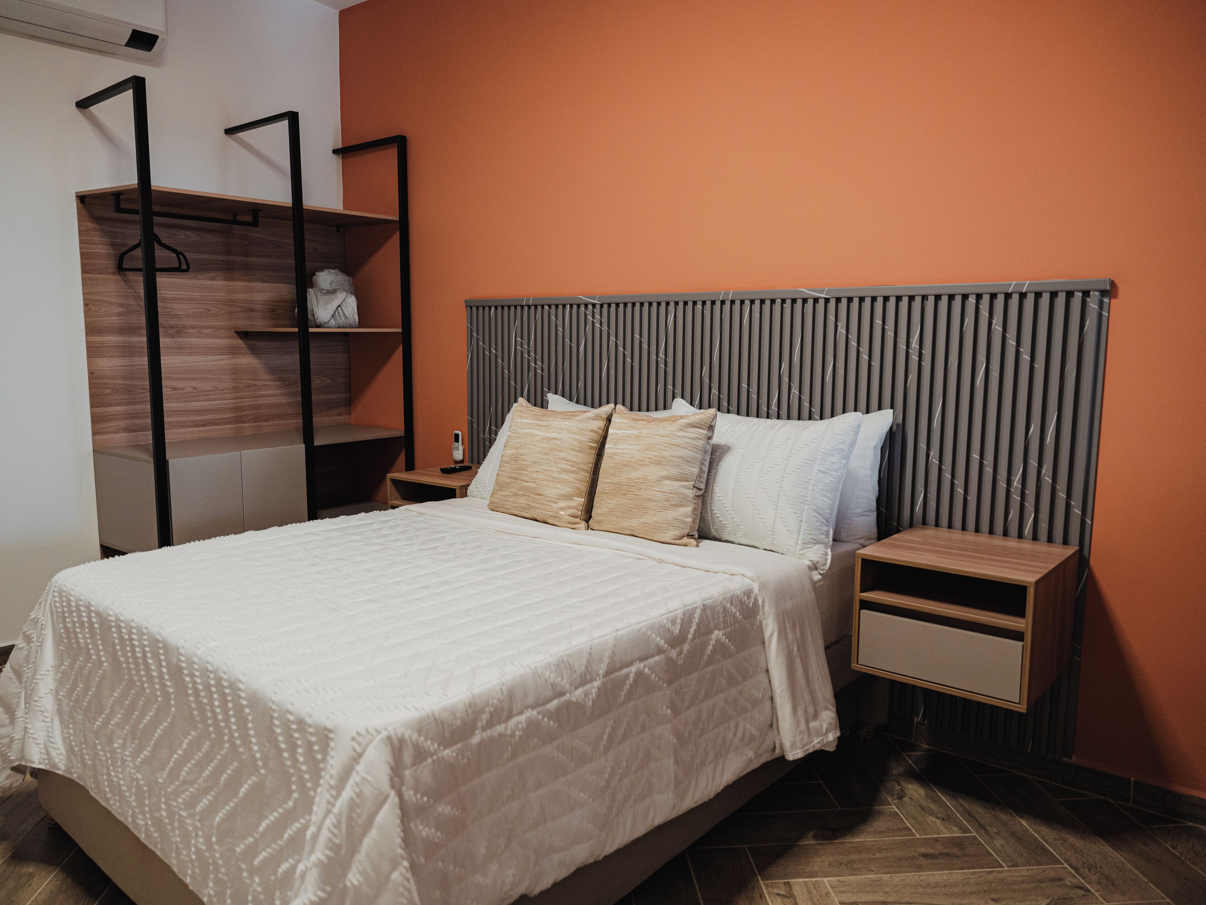 A neatly made bed with a white quilt, a wooden nightstand with a drawer, and a shelving unit with a hanging rack.