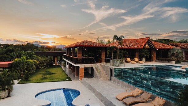 A luxurious resort with a large swimming pool, a covered outdoor area with lounge chairs, and a view of the sunset.