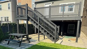 Un patio trasero con una escalera de madera que lleva a una terraza, una mesa de picnic y un área de patio cubierta.