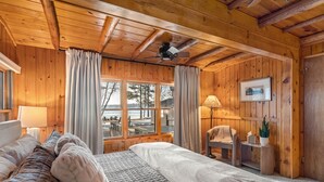 Un dormitorio de cabaña de madera con una cama, una silla, una mesita de noche, una ventana con vista a la playa y un ventilador de techo.