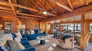 Interior espacioso de una cabaña de madera con techo alto, una zona de estar central con sofás y una mesa de centro, y un comedor con vistas al exterior.
