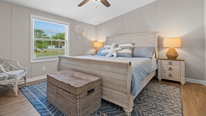 A bedroom with a wooden bed, a ceiling fan, a nightstand with a lamp, a wicker basket, and a window with blinds. - Coastal 3BR w/Game Room, 5 mins to beach, FirePit (Shallotte)