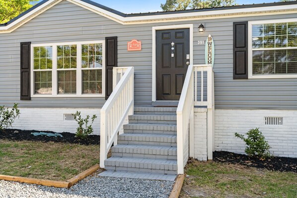 A single-story house with a dark front door, a 'No Entry' sign, and a 'Welcome' sign. There are steps leading to the door, and a small garden area with gravel and plants. - Coastal 3BR w/Game Room, 5 mins to beach, FirePit (Shallotte)