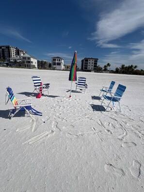 Ein Strand mit mehreren gestreiften Liegestühlen und einem farbenfrohen Sonnenschirm, im Hintergrund sind Gebäude zu sehen.