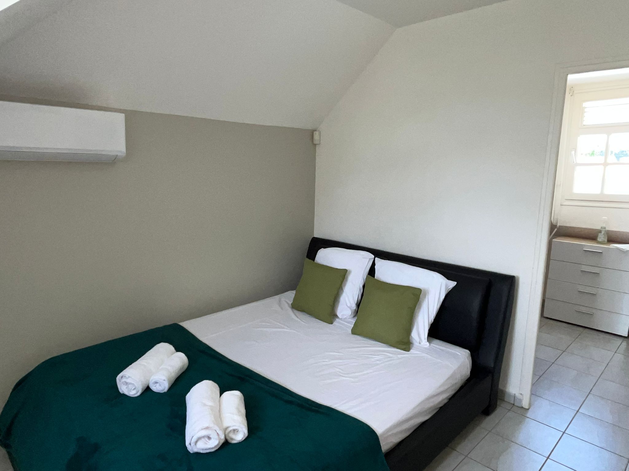 A neatly made bed with white linens and green pillows, a green throw blanket, and rolled towels on the bed.