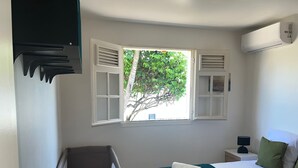 A bedroom with a bed, a nightstand, a lamp, a window with shutters, and an air conditioning unit. - Villa LOMA, overlooking lush garden and private pool. Ideal for 8 people. (Sainte-Rose)