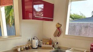 A kitchen with red cabinets, a sink, a toaster, and a window with a view of palm trees. - Villa LOMA, overlooking lush garden and private pool. Ideal for 8 people. (Sainte-Rose)