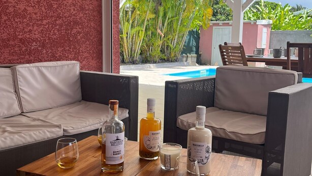 A cozy outdoor seating area with a pool view, featuring a wooden coffee table with bottles and glasses. - Villa LOMA, overlooking lush garden and private pool. Ideal for 8 people. (Sainte-Rose)