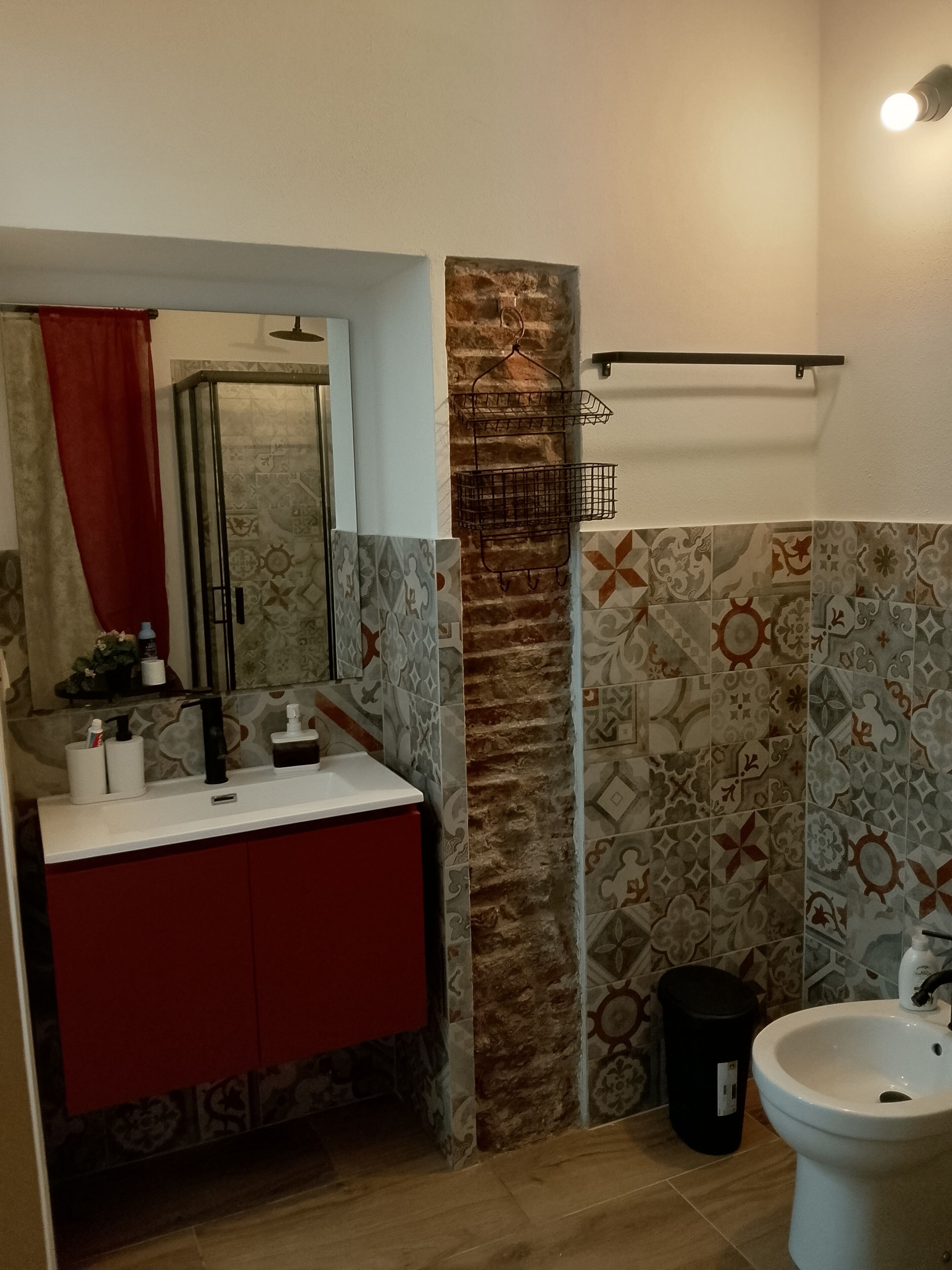 A bathroom with a sink, mirror, and toilet. There is a red cabinet under the sink and a black trash bin next to the toilet.