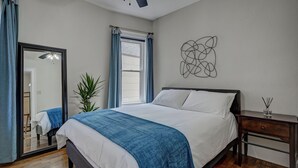 Une chambre à coucher avec un grand lit, une table de chevet, un miroir, une fenêtre avec des rideaux et une œuvre d’art murale.