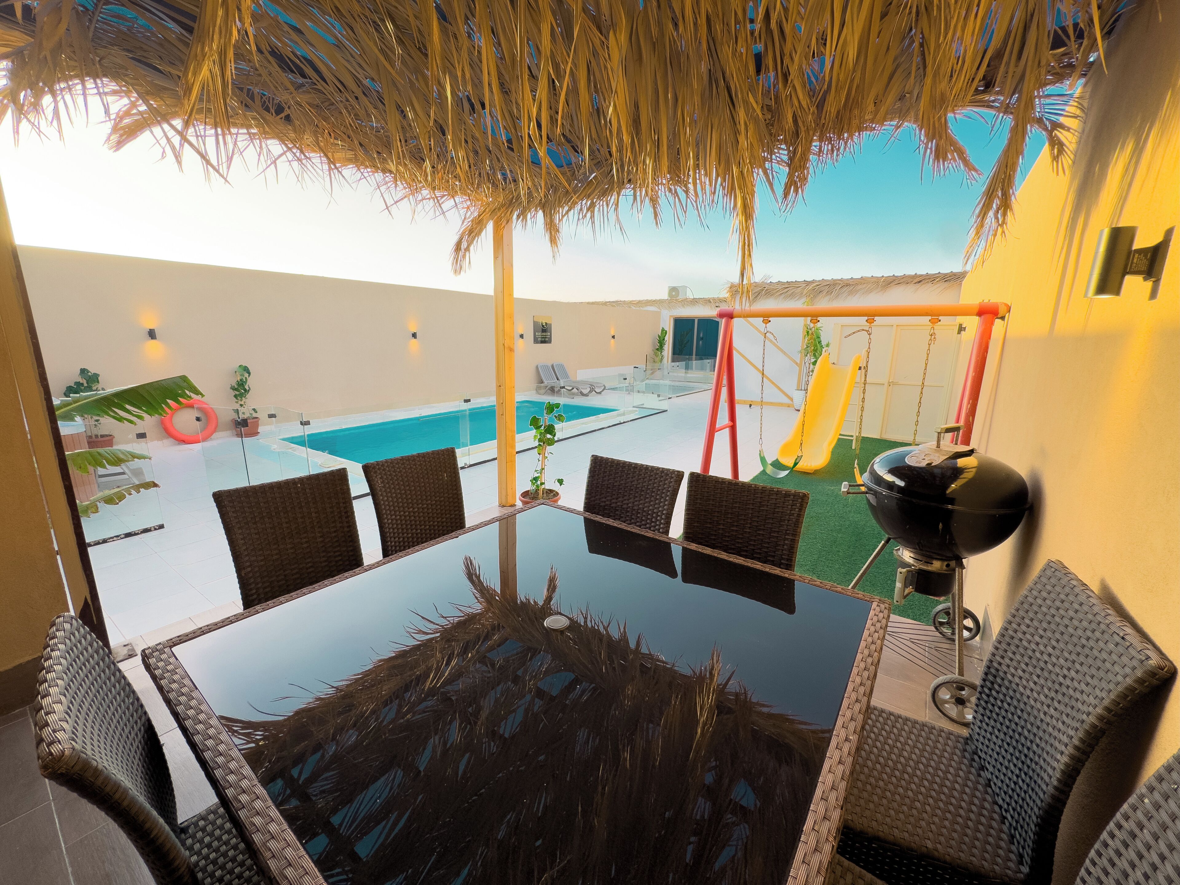 A glass-top table with wicker chairs, a pool, a swing set, and a slide.