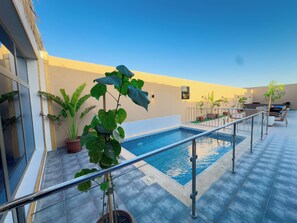 Zona de piscina en la azotea con barandilla de vidrio, plantas en macetas y un cartel con el nombre del establecimiento.
