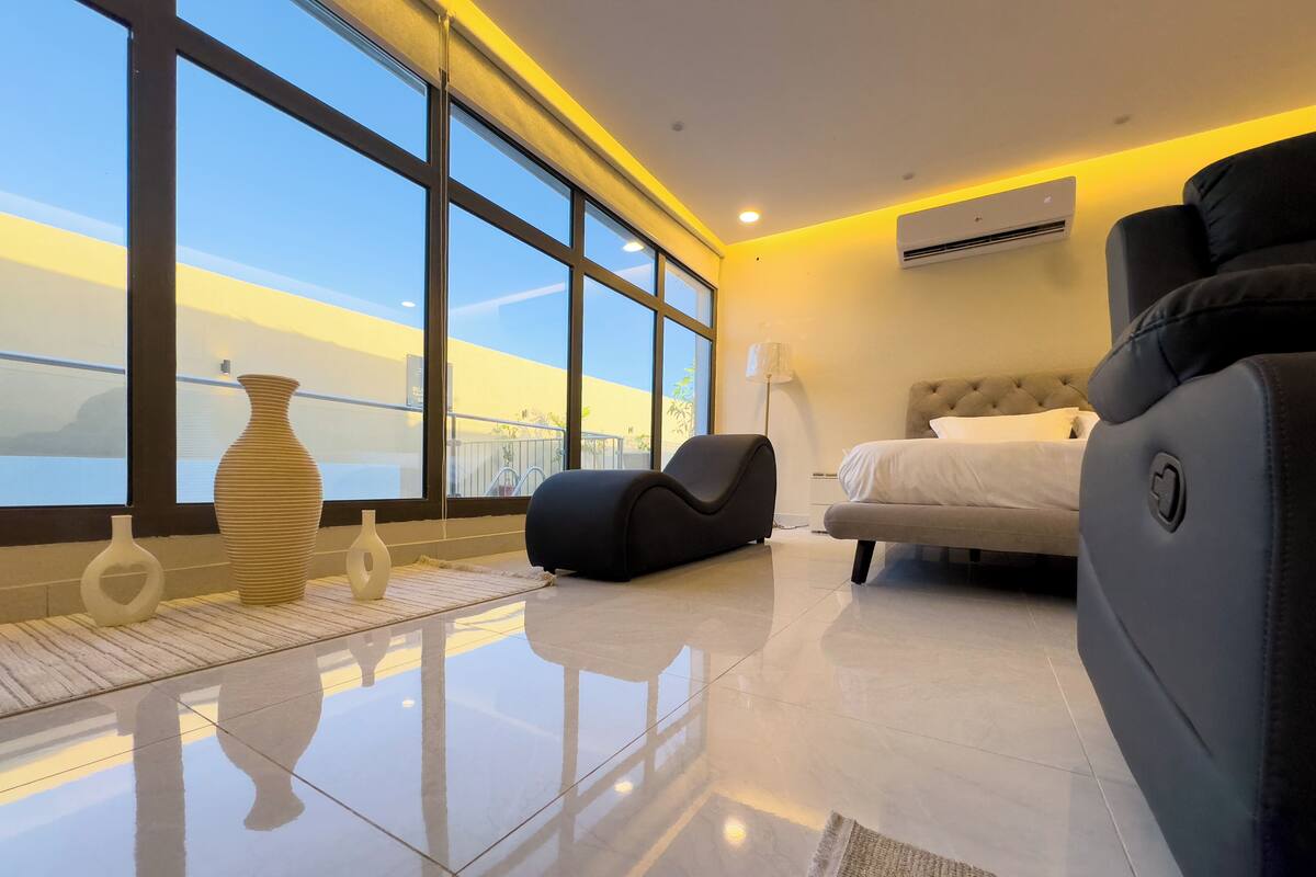 A modern hotel room with large windows, a bed, a sofa, and decorative vases.