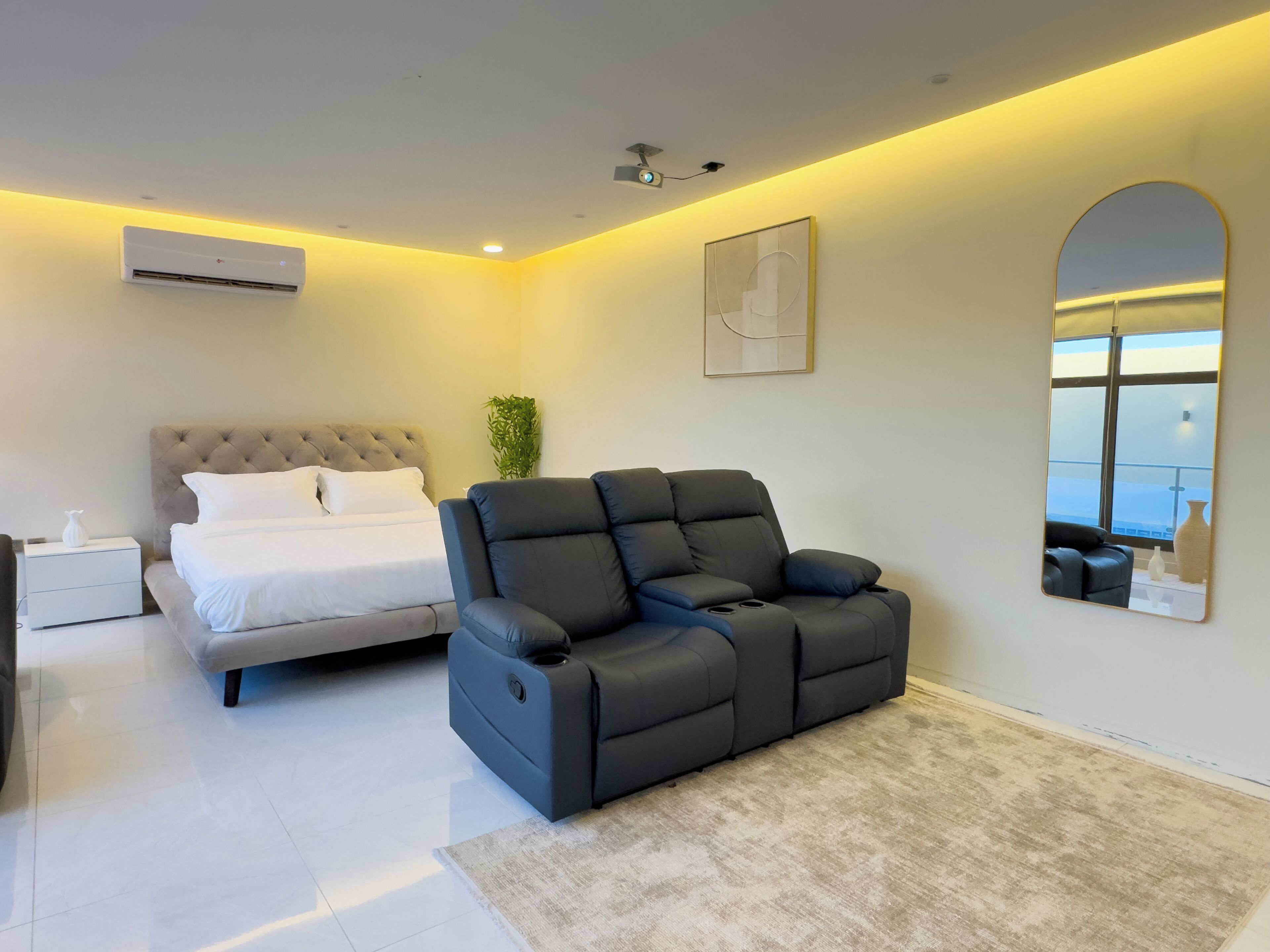 A modern bedroom with a bed, a reclining chair, a bedside table, a wall-mounted air conditioner, a ceiling projector, and a large mirror reflecting a view.