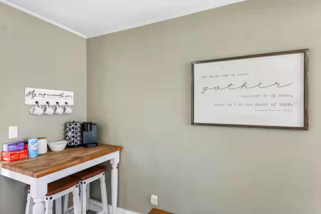 A kitchen with a wooden table, white bar stools, and a wall-mounted coffee machine. There are several mugs hanging on the wall and a framed quote above the table.