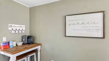 A kitchen with a wooden table, white bar stools, and a wall-mounted coffee machine. There are several mugs hanging on the wall and a framed quote above the table.