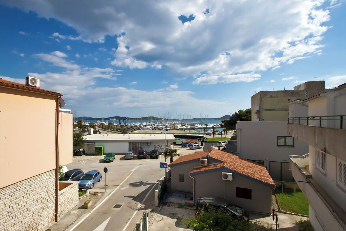 A parking lot with several cars, a building with a tiled roof, and a marina in the background.