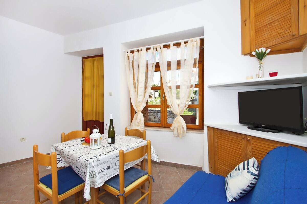 A small, cozy room with a dining table, chairs, a sofa, a television, and a window with curtains.