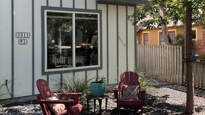 Een tweelaags huis met een groot raam en een deur, een klein terras met twee Adirondack-stoelen en een boom op de voorgrond.