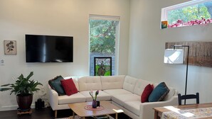 Een moderne woonkamer met een hoekbank, een salontafel, een flatscreen televisie, een plafondventilator en een groot raam met uitzicht op bomen.