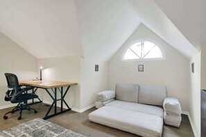 Ein Loft-Zimmer mit einem Holztisch, einem schwarzen Bürostuhl, einem grauen Sofa und einem kleinen Fenster.