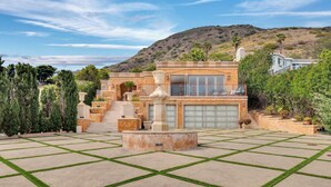 Una casa grande y moderna con un patio amplio, una fuente y montañas al fondo.