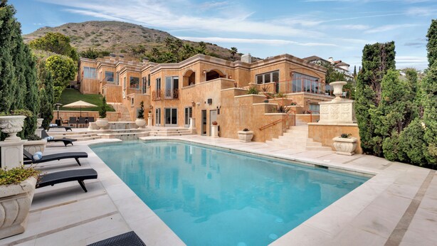 Un área de piscina lujosa con una gran piscina, tumbonas y una casa señorial con varios balcones y un paisaje de montañas al fondo.