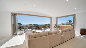 Amplia sala de estar con un sofá seccional beige grande, un comedor con mobiliario de exterior y ventanas amplias que ofrecen vistas al mar y follaje.