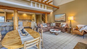 A spacious living area with a wooden dining table, a brown leather sofa, and a coffee table. There is a kitchen in the background with wooden cabinets and a bar area. - Ski-in/out spacious 3BR Saddle Ridge townhome, steps to Ironhorse Lift & hot tub (Big Sky)