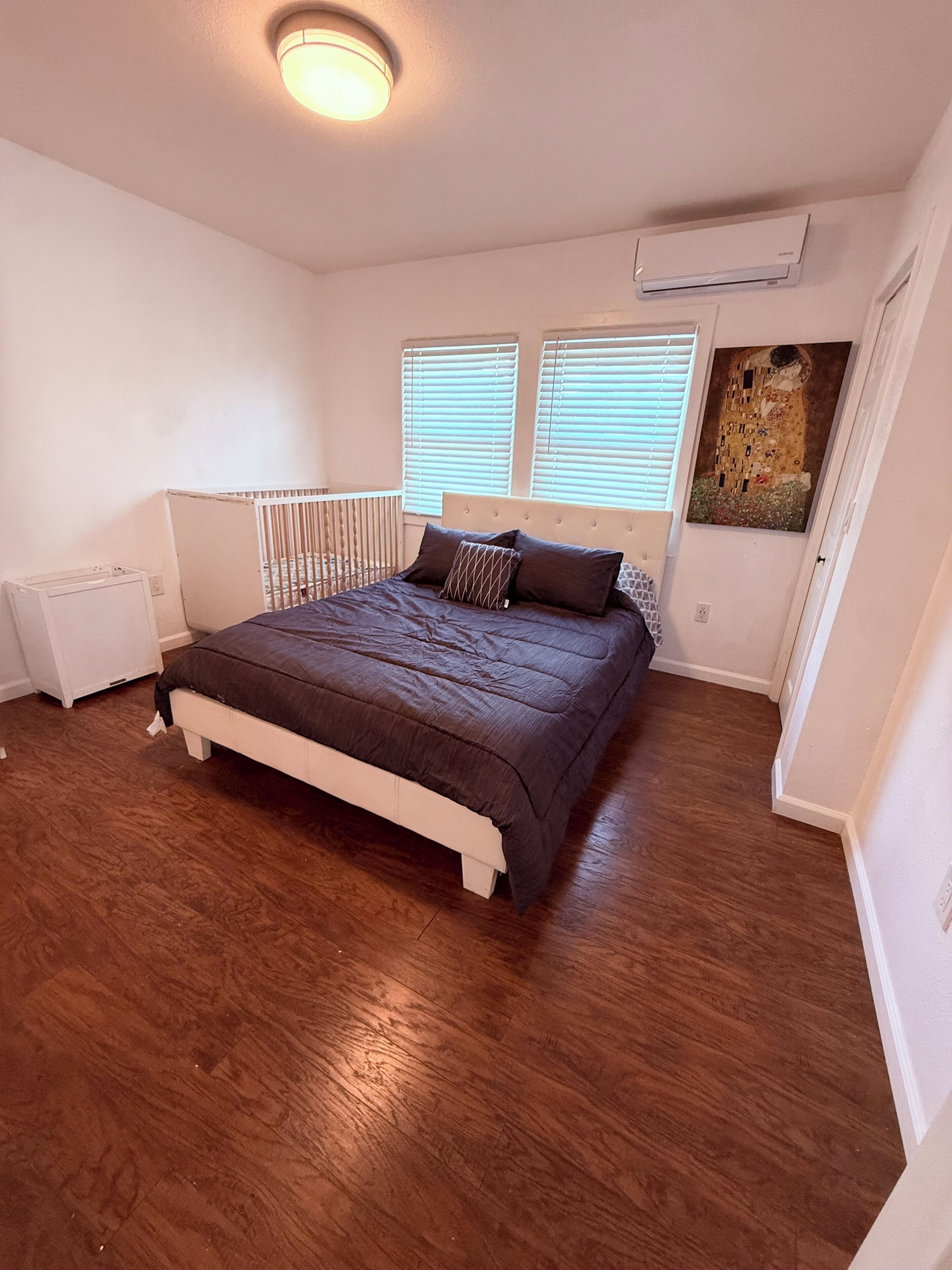 Una camera da letto con un letto, pareti bianche, una lampada a soffitto, una finestra con persiane e un quadro appeso al muro.