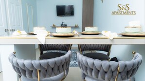 A dining area with a white table, four grey upholstered chairs with gold legs, and a television mounted on the wall.