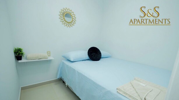 A bedroom with a single bed, a small shelf with a plant and a towel, a round mirror, and a sunburst wall decoration.