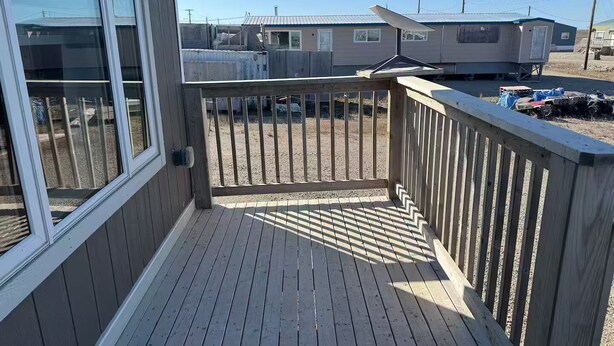 Una terraza de madera con baranda metálica, vista despejada a un patio de grava y una hilera de casas móviles al fondo.