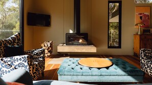 A living room with a wood-burning stove, patterned sofas, a television, and a large window with a view of trees. - Parachilna at Boardwalk Villas Metung (Metung)