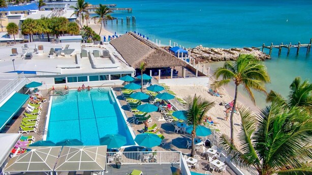 Un resort con una grande piscina, lettini prendisole e una spiaggia attrezzata con ombrelloni.