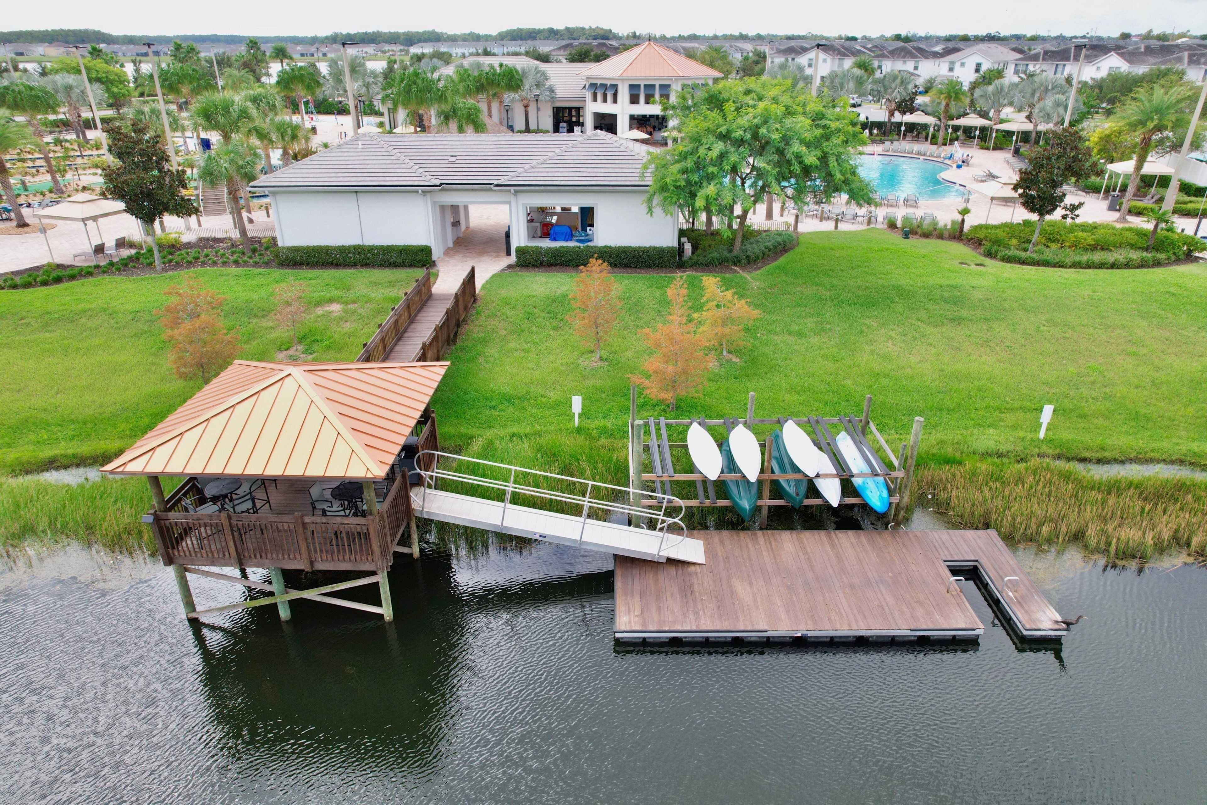 A waterfront property with a wooden dock, a covered seating area, and a pool in the background.