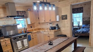 Un intérieur en bois comprenant un coin cuisine, une table à manger avec des bancs et un lit recouvert d’une courtepointe à motifs.
