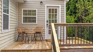 A wooden deck with two wicker chairs and a small table, a door with a window, and a wooden railing. - The Blue Door Bungalow. Walk to Duke Medical/VA. (Durham)