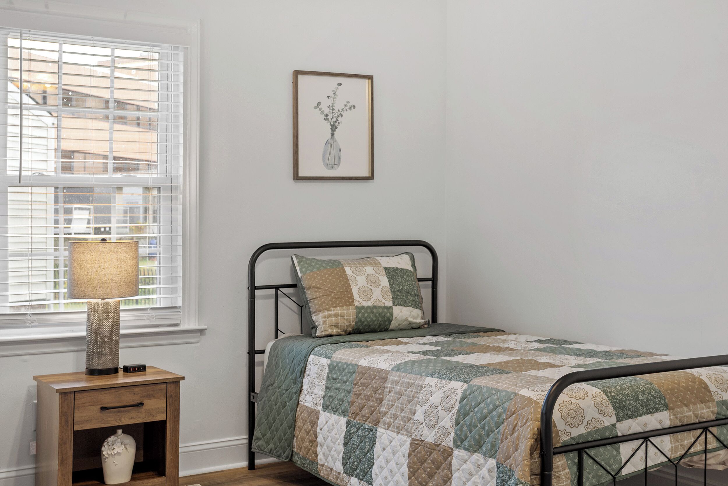 A neatly made bed with a patchwork quilt, a bedside lamp, a small nightstand with a vase, and a framed picture on the wall.