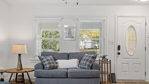 A living room with a grey sofa, a round wooden side table, a floor lamp, and a white door with a stained glass window. - The Blue Door Bungalow. Walk to Duke Medical/VA. (Durham)